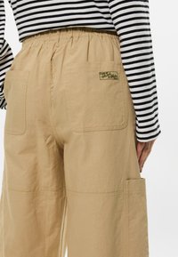 Καφέcargo παντελόνι με ελαστική μέση, διαθέτοντας δύο πλαινές τσέπες και μία πίσω τσέπη με πράσινη ετικέτα. Ριγέ μαύρη και λευκή μπλούζα.