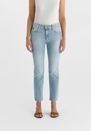 Vrouw draagt lichtblauwe slim-fit jeans, wit mouwloos topje en bruine open teen hakken, staand tegen een effen witte achtergrond.