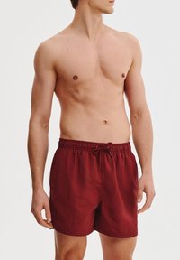 Czerwone kąpielówki wykonane z lekkiego materiału, z elastycznym pasem i sznurkiem do regulacji, w luźnym kroju. Brak widocznych wzorów lub elementów metalowych.