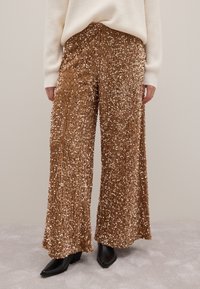 Goldene, paillettenbesetzte weite Hosen mit einer strukturierten Oberfläche, kombiniert mit einem weißen Strickpullover und schwarzen, absatzhohen Stiefeletten.