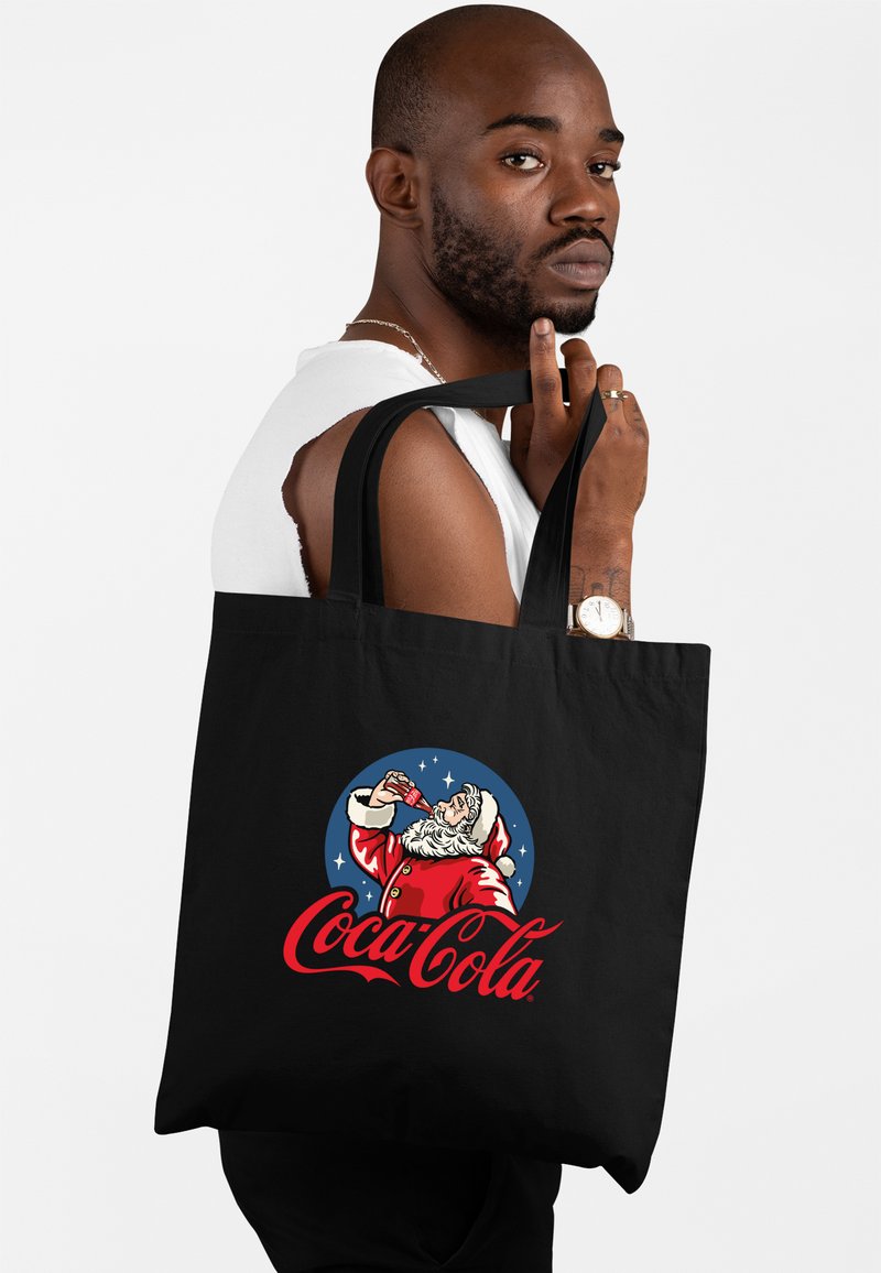 Czarna torba typu tote z grafiką świętego Mikołaja Coca-Cola w stylu retro, z żywymi kolorami i teksturowanymi elementami designu. Regulowane materiałowe paski.