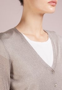 Ljusgrå, metallisk cardigan med V-ringning, som har två knappar och genomskinliga ärmar, lager på lager över en vit T-shirt.