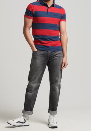 Mann trägt ein rot-navy gestreiftes Polohemd, graue umgeschlagene Jeans und weiße Turnschuhe, steht mit einer Hand in der Tasche vor einem einfarbigen Hintergrund.