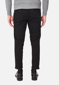 Pantaloni di cotone neri con orli arrotolati, due tasche posteriori con bottoni, abbinati a scarpe nere lucide, visti da dietro.