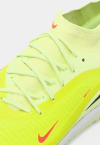 Botín de fútbol amarillo de Nike con logotipos en forma de swoosh rojos, cordones verde claro y un cuello de calceta, vista de cerca que muestra los detalles.