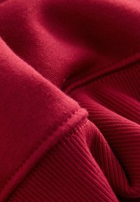 Punainen kangas, jossa on sileä pinta ja uritetut korostukset. Materiaali on pehmeä, ja siinä on näkyviä ompeleita, jotka korostavat sävyjen ja taitteiden vaihteluita.