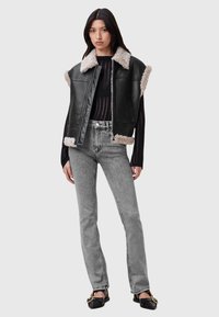 Gilet di pelle nera con bordo in pelliccia sintetica, abbinato a jeans grigi chiari a vita alta. Il modello è in piedi su uno sfondo neutro.