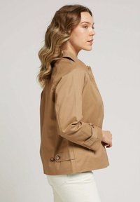 Veste légère beige au design structuré, dotée d'un col, de détails en boutons sur les poignets et d'une texture lisse et mate.