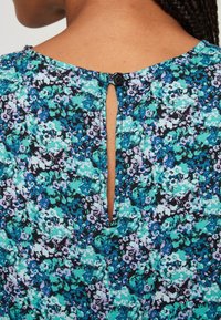 Blommig blus med ett mönster i svart, turkos och lila; har en rund halsringning med en nyckelhålsöppning och en svart knappknäppning bak.