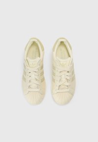 Ténis cor creme com biqueira de borracha texturizada. Apresentam três riscas verde claro, colarinho almofadado e atacadores planos. Design minimalista.