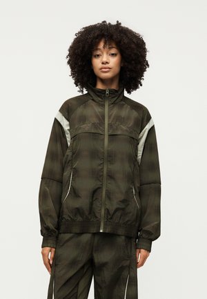 Γυναίκα με σγουρά μαλλιά φοράει σκούρο πράσινο καρό μπουφάν με φερμουάρ και παντελόνια που ταιριάζουν, μπροστά σε απλό φωτεινό φόντο.