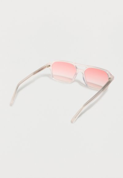 Γυαλιά ηλίου με διαφανές πλαίσιο και τετράγωνα φακούς με ροζ διαβάθμιση, ανοιχτά πάνω σε λευκή επιφάνεια.