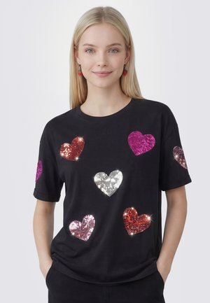 Donna bionda che indossa una maglietta nera con cuori di paillettes multicolori lucenti e orecchini a forma di fragola. Mani in tasca.