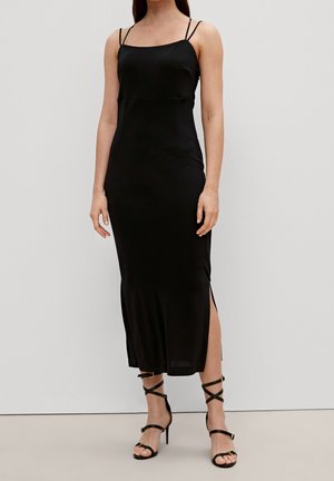 Woman wearing a sleeveless black midi dress with thin straps and side slit, paired with black strappy high-heeled sandals.