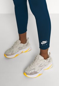 Nike Air sneakers i beige med grå accenter och en gul sula, utrustade med meshpaneler och en framträdande logotyp på sidan. Bärs med mörkblå leggings.