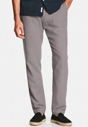 Man draagt grijze rechte broek met zwarte slip-on espadrilles en een donker overhemd, staat met één hand in de zak.