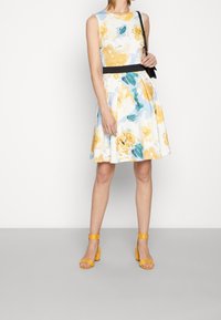 Vestido de estampado floral en colores pastel con cintura entallada y falda acampanada. Combinado con tacones amarillos de tiras al tobillo. Incluye un bolso negro.