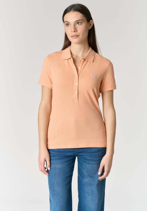 Donna con lunghi capelli scuri che indossa una polo color pesca a maniche corte e jeans blu, in piedi contro uno sfondo chiaro e uniforme.