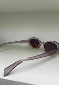 Lunettes de soleil ovales avec des montures transparentes de couleur rose clair et des verres foncés. Branches fines et droites avec un logo d'un côté.