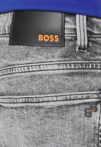 Šedé džíny z denimu s černou koženou nášivkou s nápisem "BOSS" v oranžové. Texturovaná tkanina s detaily šití a černou logovou akcentací.