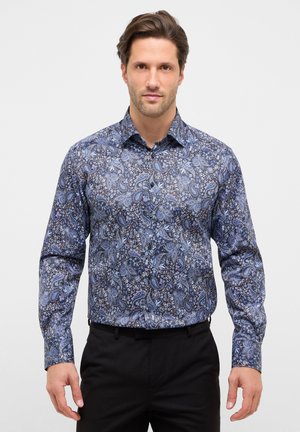 Uomo che indossa una camicia a maniche lunghe con motivo paisley blu infilata in pantaloni neri, in piedi davanti a uno sfondo bianco semplice.
