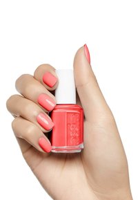 Vernis à ongles corail dans un flacon en verre, tenu dans une main avec des ongles corail mates assortis. Le flacon est doté d'un bouchon blanc et d'un logo en relief.