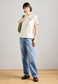 Ανοιχτό μπεζ βαμβακερό t-shirt με σχέδιο γραφικών ματιών, σε συνδυασμό με φαρδιές μπλε τζιν παντελόνες με καμπύλες ραφές, και καφέ παπούτσια με χοντρό τακούνι.