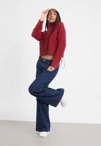 Rode gebreide cropped trui met ruitpatronen, gecombineerd met donkerblauwe flared jeans en witte sneakers. Het model draagt een witte pet.