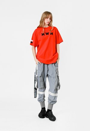 Camiseta de gran tamaño roja con el logo "MWM"; pantalones cargo grises con múltiples bolsillos y detalles de cremallera; calzado negro texturizado.