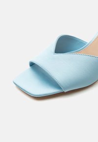 Ανοιχτή μπεζ σανδάλι με τετράγωνη μύτη σε ανοιχτό μπλε χρώμα, με φαρδιά υφασμάτινη λωρίδα και εσωτερική σόλα σε μπεζ απόχρωση, πάνω σε λευκό φόντο.