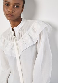 Weiße Bluse aus leichtem Stoff, mit einem Rüschensattel, floralem Stickereimuster und einer durchgehenden Knopfleiste. Lange Ärmel inklusive.