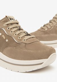 Baskets en daim beige avec semelles blanches épaisses, lacets beige, œillets dorés, perles métalliques décoratives sur les lacets et logo en relief sur le côté.