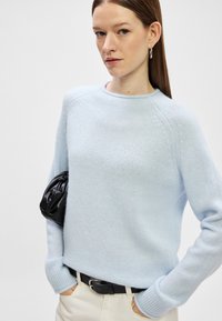 Světle modrý pletený svetr s dlouhými rukávy, žebrovanými manžetami a kulatým výstřihem. Kombinován s černou psaníčkem a světlými kalhotami.