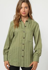 Camisa verde oliva de botones con rayas verticales, mangas largas y bolsillo en el pecho. Presenta botones marrones y un diseño de corte relajado.