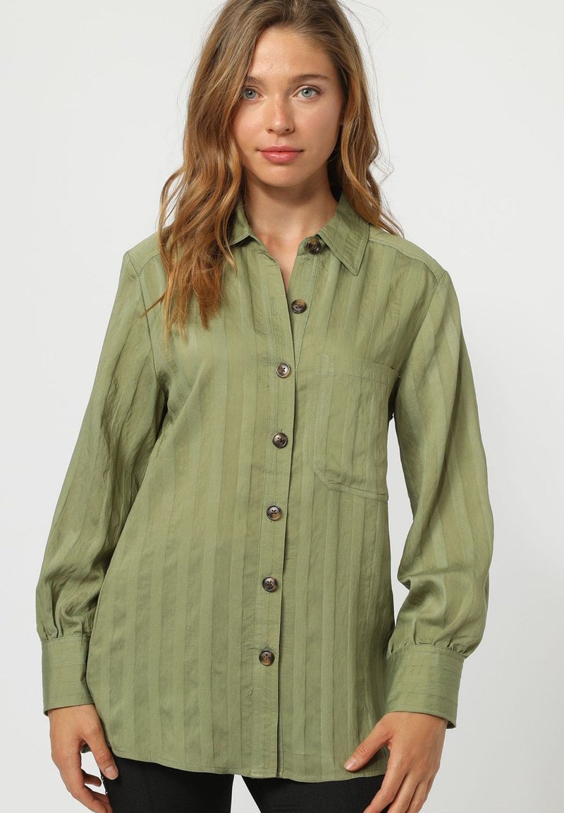 Camisa verde oliva de botones con rayas verticales, mangas largas y bolsillo en el pecho. Presenta botones marrones y un diseño de corte relajado.