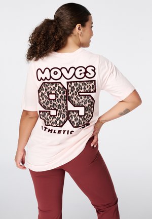Vrouw met krullend haar in een paardenstaart draagt een lichtroze shirt met "Moves 95" in luipaardprint op de rug en bordeaux leggings, staat met hand op de heup.