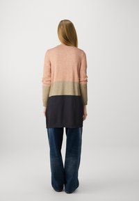 O suéter longo apresenta um design em bloco de cores em pêssego, bege e azul-marinho escuro, com um corte descontraído e punhos canelados. Combinado com jeans de perna larga.