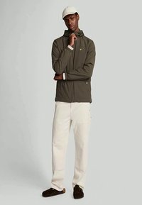 Young man in olive green hooded jacket, white pants, dark shoes, and white cap standing with one hand on chin and the other arm crossed.