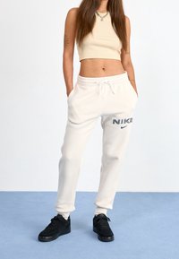 Valkoiset puuvillahousut, joissa on joustava vyötärönauha ja nyöri, sekä vasemmassa reidessä painettu musta "NIKE" -logo. Mustat lenkkarit täydentävät asua.