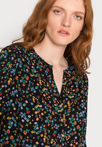 Blusa preta de flores com decote em V, padrões florais coloridos em vermelho, azul e amarelo, e botões decorativos na parte da frente.
