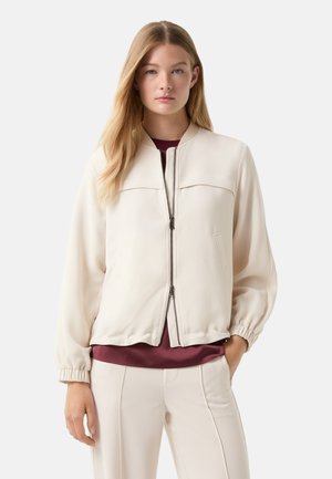 Veste bomber crème avec une texture lisse, fermeture éclair, deux poches avant et des poignets froncés, portée avec un haut bordeaux en dessous.