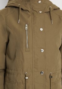 Khaki zip-up jacka med huva, utrustad med silverfärgad hårdvara, en ficka med dragkedja fram och justerbar dragsko vid midjan. Slät textur.