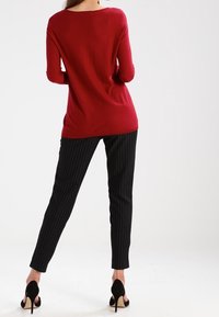 Haut rouge à manches longues avec un large décolleté, associé à un pantalon noir à fines rayures. Chaussures à talons hauts noirs. Design simple, texture douce.