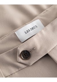 Beżowa tkanina o teksturowanej powierzchni z widocznym przeszyciem. Zawiera okrągły, ciemny guzik oraz białą metkę z napisem "LES DEUX".