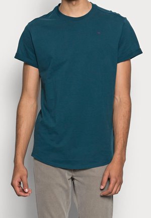Homme portant un t-shirt col rond à manches courtes de couleur bleu sarcelle et un pantalon gris clair, debout avec les bras détendus devant un fond uni.