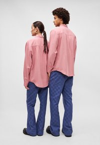 Duas pessoas estão de costas uma para a outra. Elas vestem camisas button-up de rosa claro e calças checkered azul-escuro, com sapatos pretos texturizados.