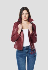 Bordová kožená bunda s vysokým golierom, prešívanými detailmi na ramenách, vreckami na zips, kombinovaná s modrými džínsami s farebnou výšivkou.