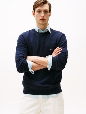 Marineblauwe sweatshirt met geribde manchetten en zoom, gedragen over een lichtblauw overhemd met kraag. Model staat met de armen gekruist tegen een witte achtergrond.