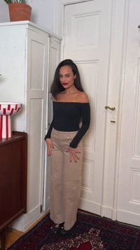 Femme aux longs cheveux foncés portant un haut noir épaules dénudées, un pantalon beige à jambes larges et des chaussures noires, debout à l’intérieur devant une porte blanche et un meuble.