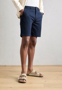 Navy cotton shorts with a tailored fit, side pockets, and a front button closure, paired with beige two-strap sandals. Natural wood flooring.